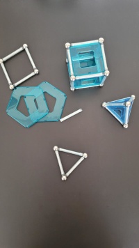 Das Bild zeigt aus magnetischen Stäben, Kugeln und Plastikteilen aus den geometrischen Grundformen Quadrat und Dreieck zusammengebaute geometrische Körper. Die Stangen und Kugeln sind silbern, die Plastikformen in einem durchsichtigem Acrylblau